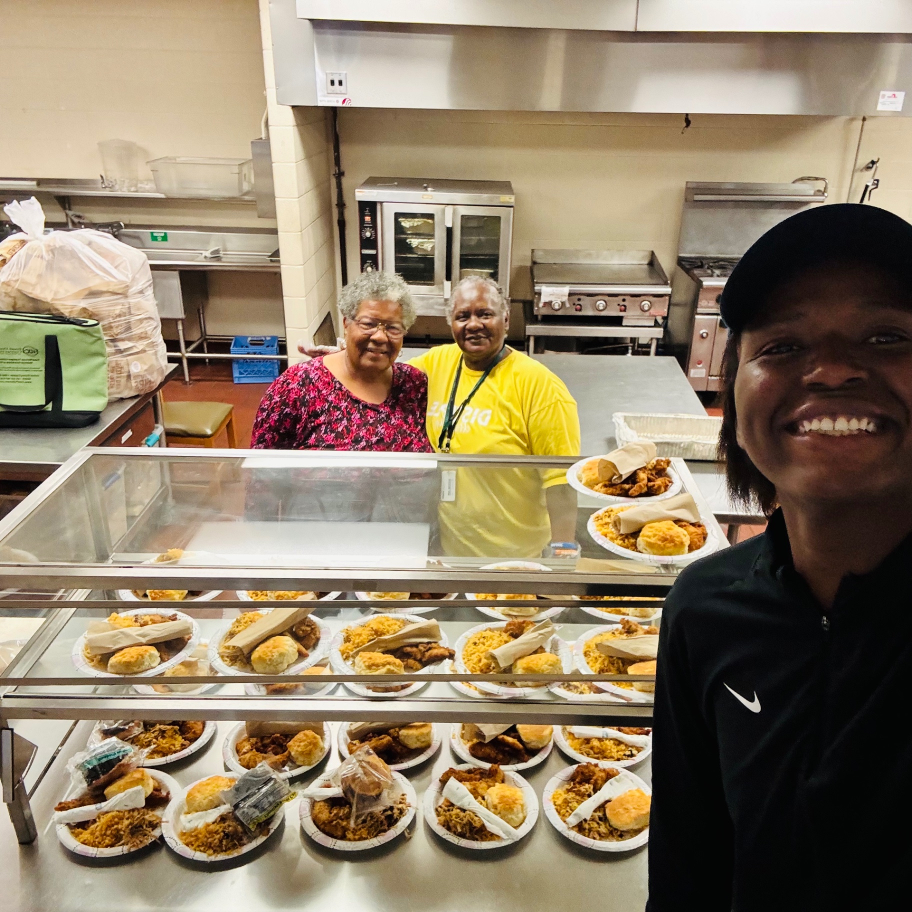 Two kitchen volunteers and Daniel smiling behind a serving counter of plated dinners